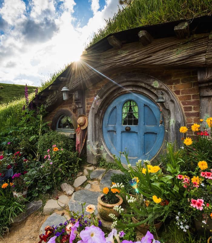 Hobbit house with blue round door and colorful garden under sunny sky with clouds.