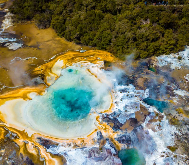 Aerial view of a colorful geothermal hot spring with steam and forest around it.
